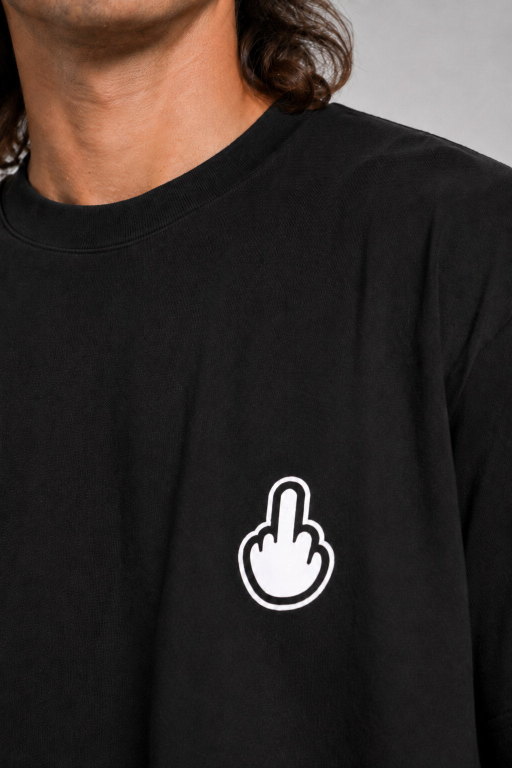 Close-up of a person wearing a black crewneck t-shirt with a white hand graphic showing the middle finger on the left chest, set against a neutral gray background. #black