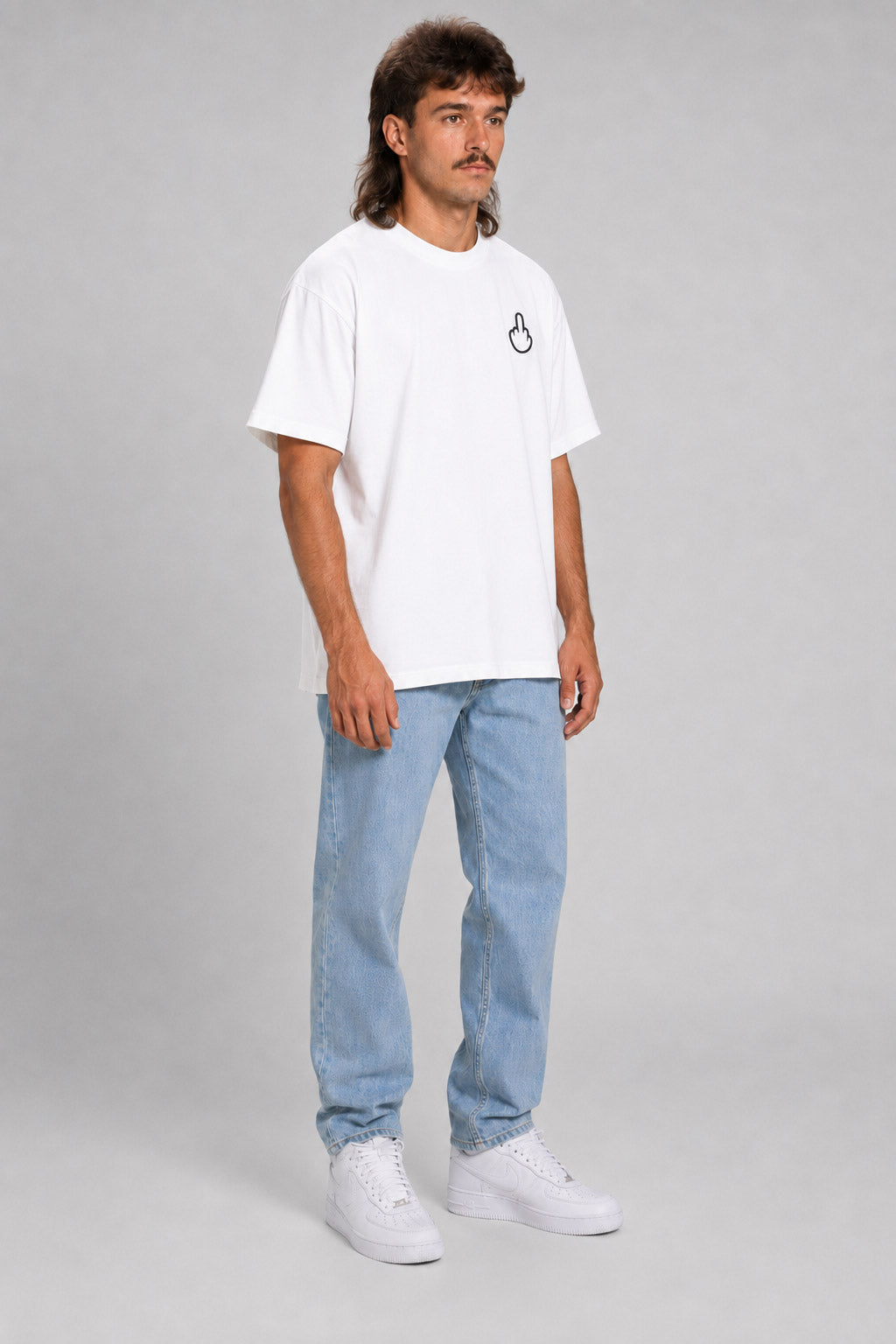 A man modeling a white short-sleeve t-shirt with a small black circular logo on the left chest, paired with light blue jeans and white sneakers, against a solid gray studio background. #white